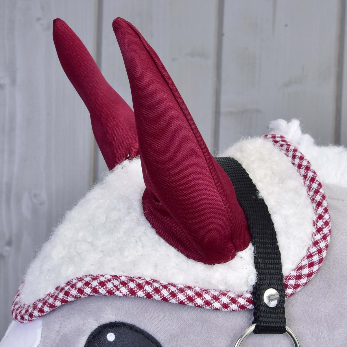 Close-up of a plush toy animal wearing the Hobby Horse LarDen Ear Bonnet Cube Red, featuring red fabric horns, checkered red-and-white trim, and a black strap with metal ring, set against a gray wooden background.