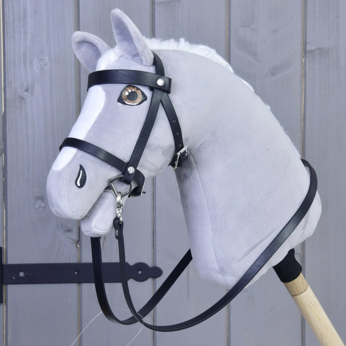 The Hobby Horse LarDen Set Basic, a gray plush hobby horse with a black bridle and reins on a wooden stick, is displayed in front of gray wooden panels.