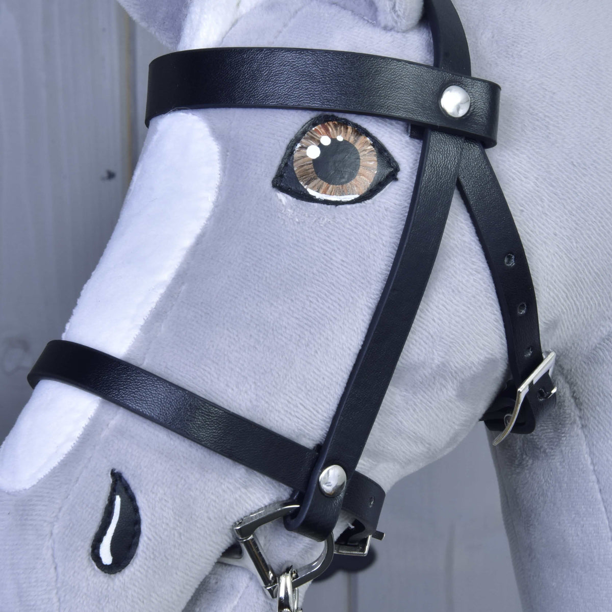 A close-up of the Hobby Horse LarDen Set Basic, a gray and white plush horse with detailed stitching, realistic embroidered eye and nostril, and a black leather bridle with reins.