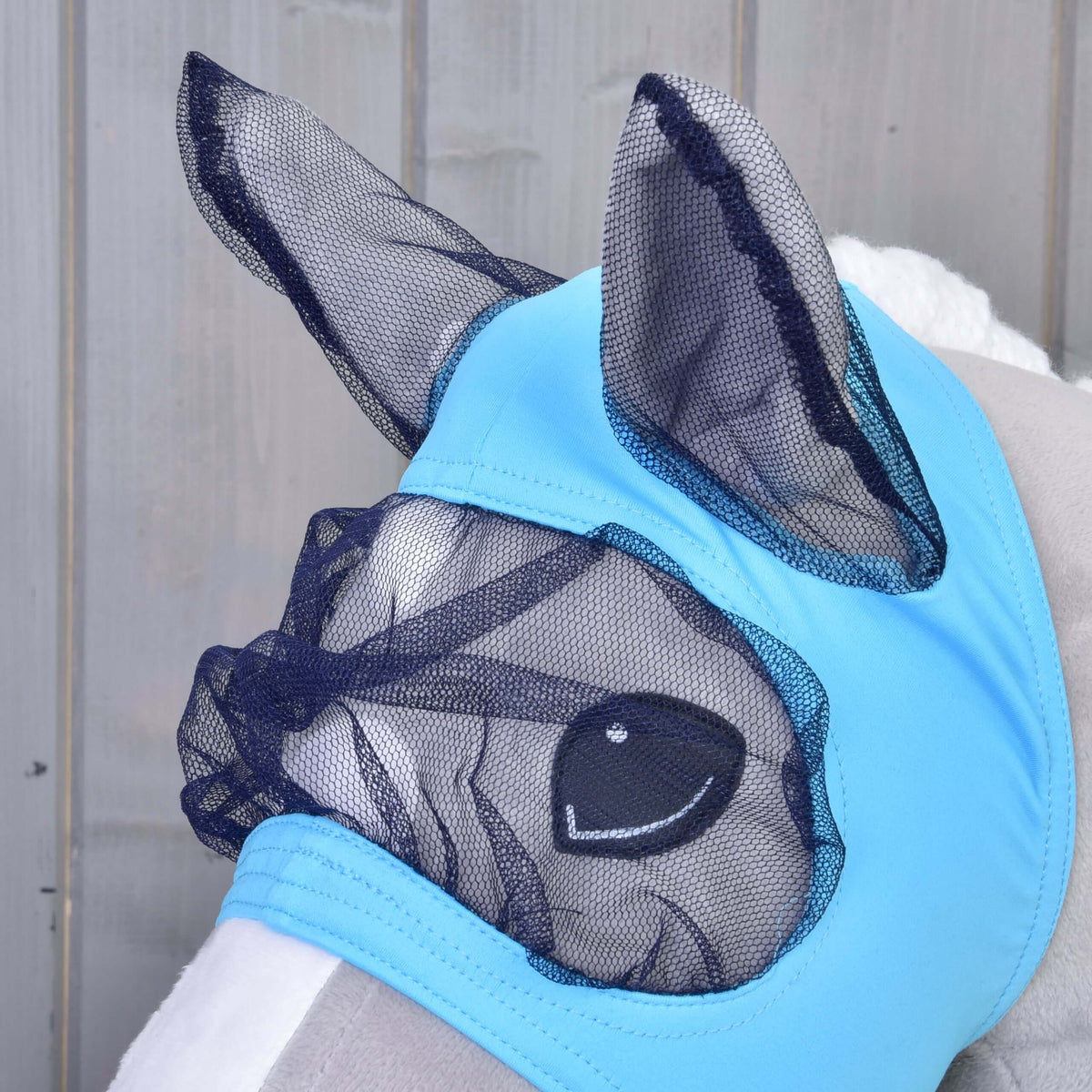 A close-up of a plush horse wearing the Hobby Horse LarDen Fly Mask, featuring light blue fabric with black mesh over its ears, eyes, and nose for protection. The washable mask is designed like those used on real horses and shown against a wooden background.