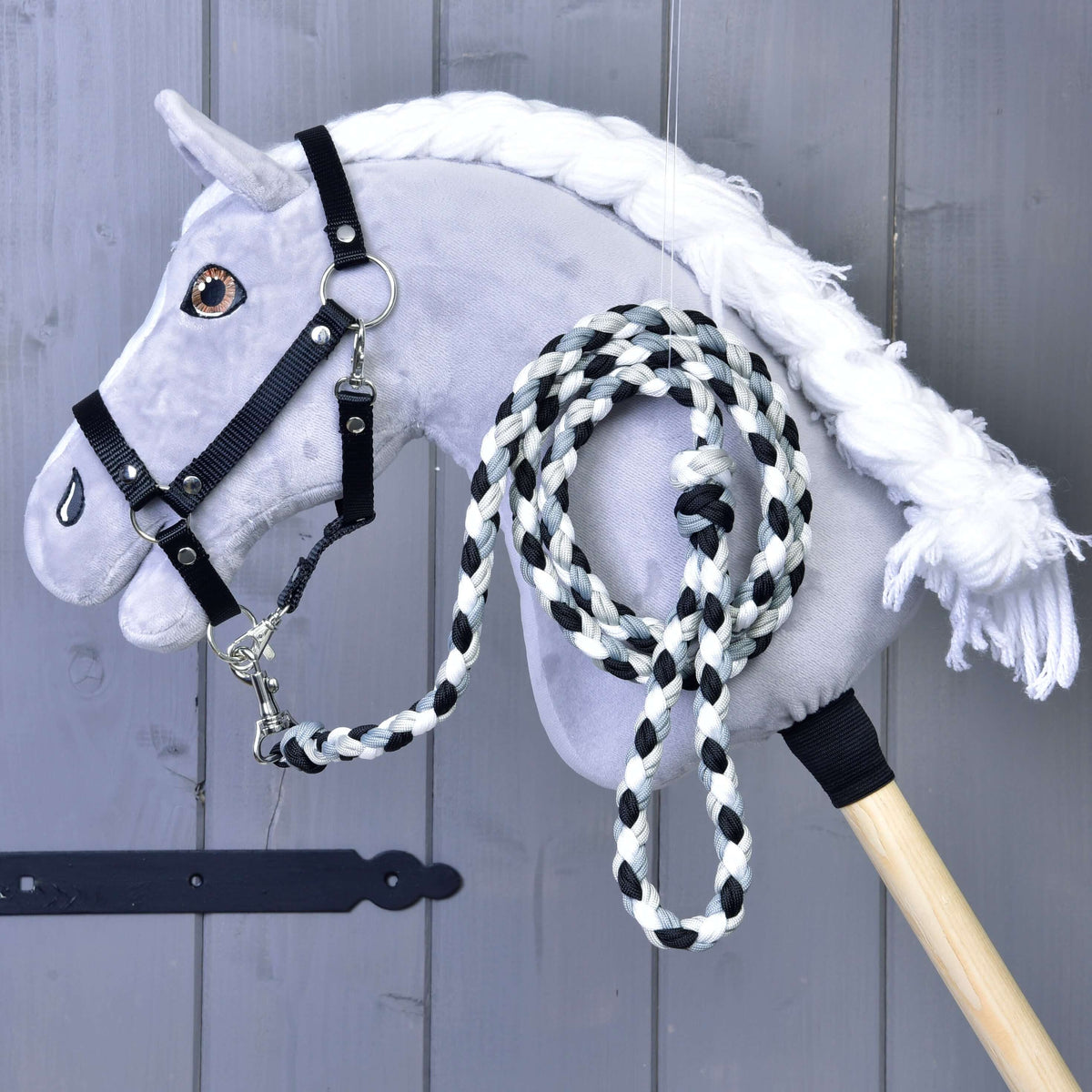 The Hobby Horse LarDen Hobby Horse features a gray plush head with a white mane, black bridle, and includes a black-and-white braided lead rope. Essential horse equipment, displayed against a gray wooden background.