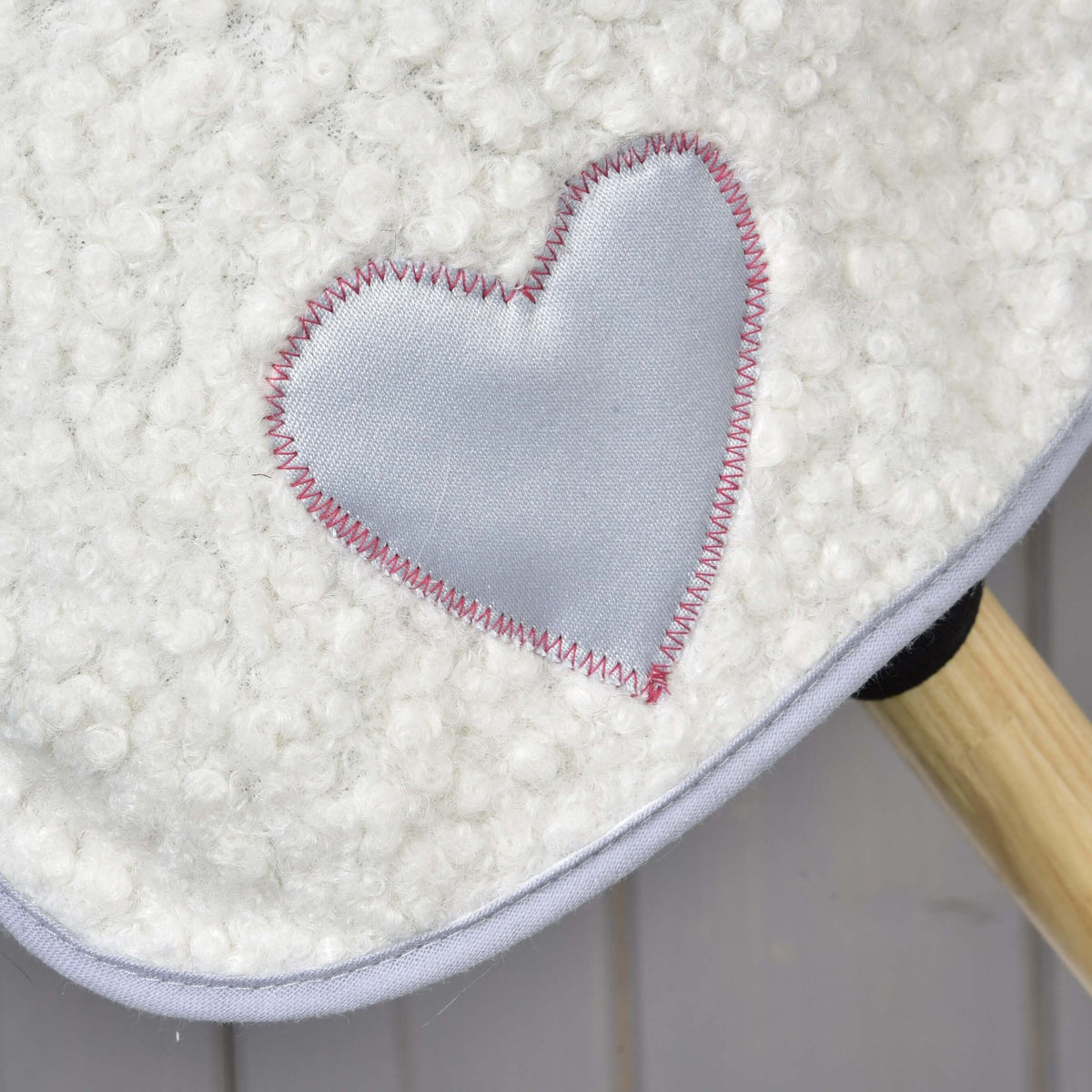 Close-up of the white textured Set Snowflake ear bonnet by Hobby Horse LarDen, featuring a shiny light gray heart patch with pink zigzag stitches; a wooden stick appears in the bottom right corner.