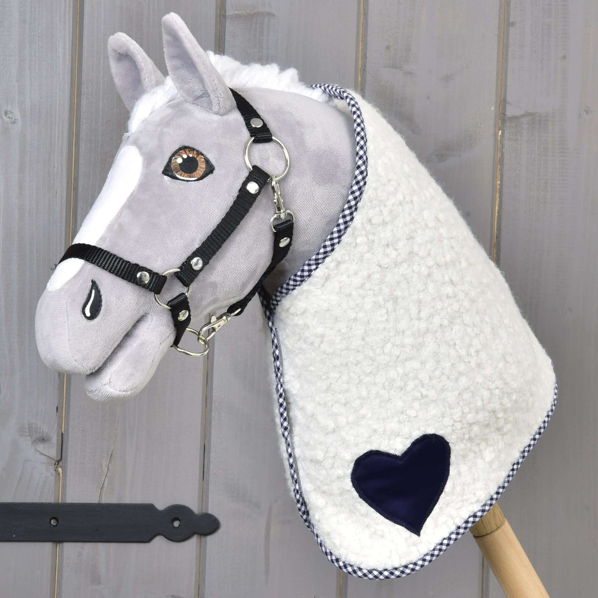The Hobby Horse LarDen "Blanket cube blue" is a plush hobby horse with a gray face, black bridle, and a blue-accented blanket, mounted on a wooden stick and set against a wooden background.