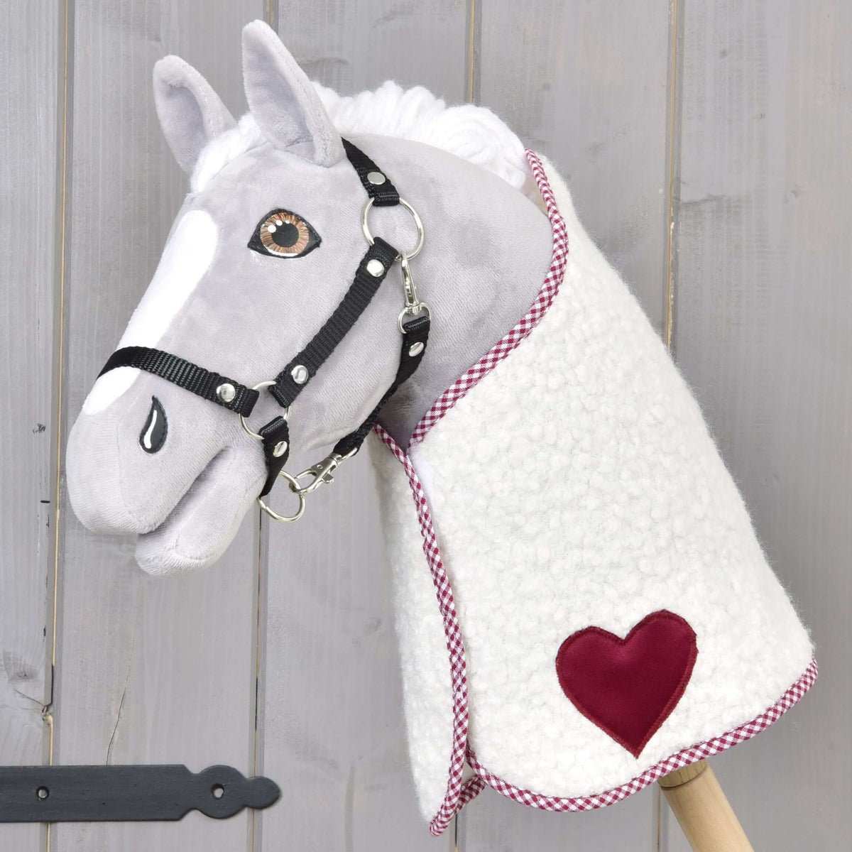 The Hobby Horse LarDen plush hobby horse has a white and gray face, black bridle, and comes with a Blanket cube red fleece blanket featuring a red heart patch, mounted on a wooden stick against light wood.
