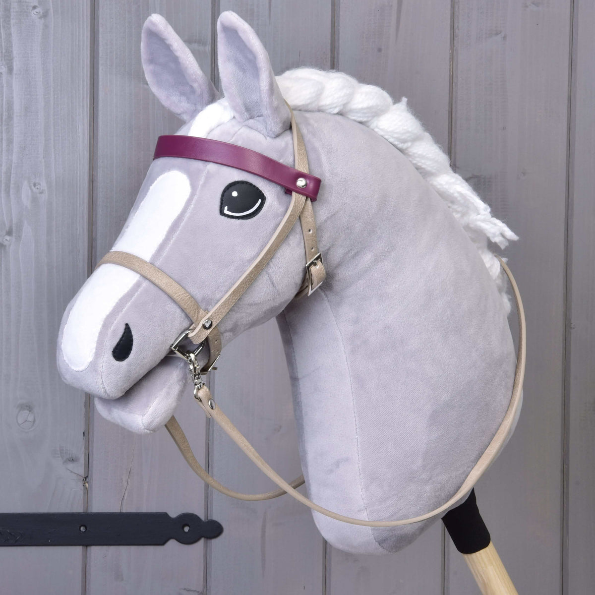 The Hobby Horse LarDen features a gray hobby horse with a white mane, black eye, and a Bordeaux leatherette set—bridle, reins, and martingale—plus a purple browband and cartoon-like face. It stands upright against a wooden background.