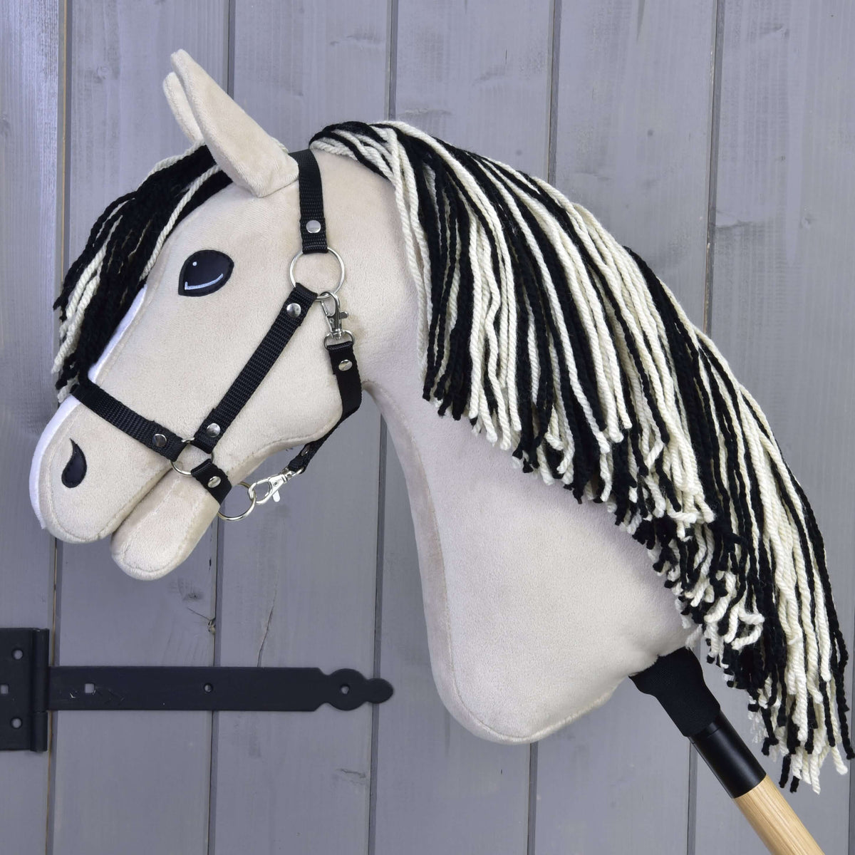 The Hobby Horse LarDen Aurora with Black Halter, featuring a beige head, black and white mane, and wooden stick, is shown against a gray wooden wall.