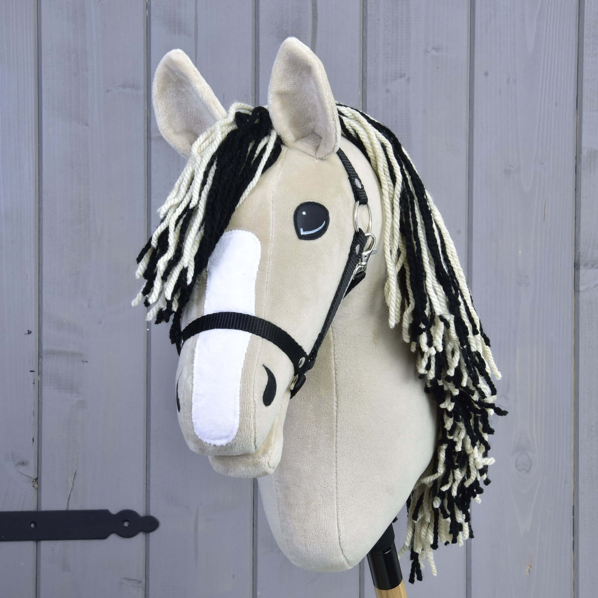 The Hobby Horse Aurora by Hobby Horse LarDen, inspired by an American Quarter Horse, features a beige body, white muzzle, black and white yarn mane, and a black halter, shown in front of a gray wooden fence.
