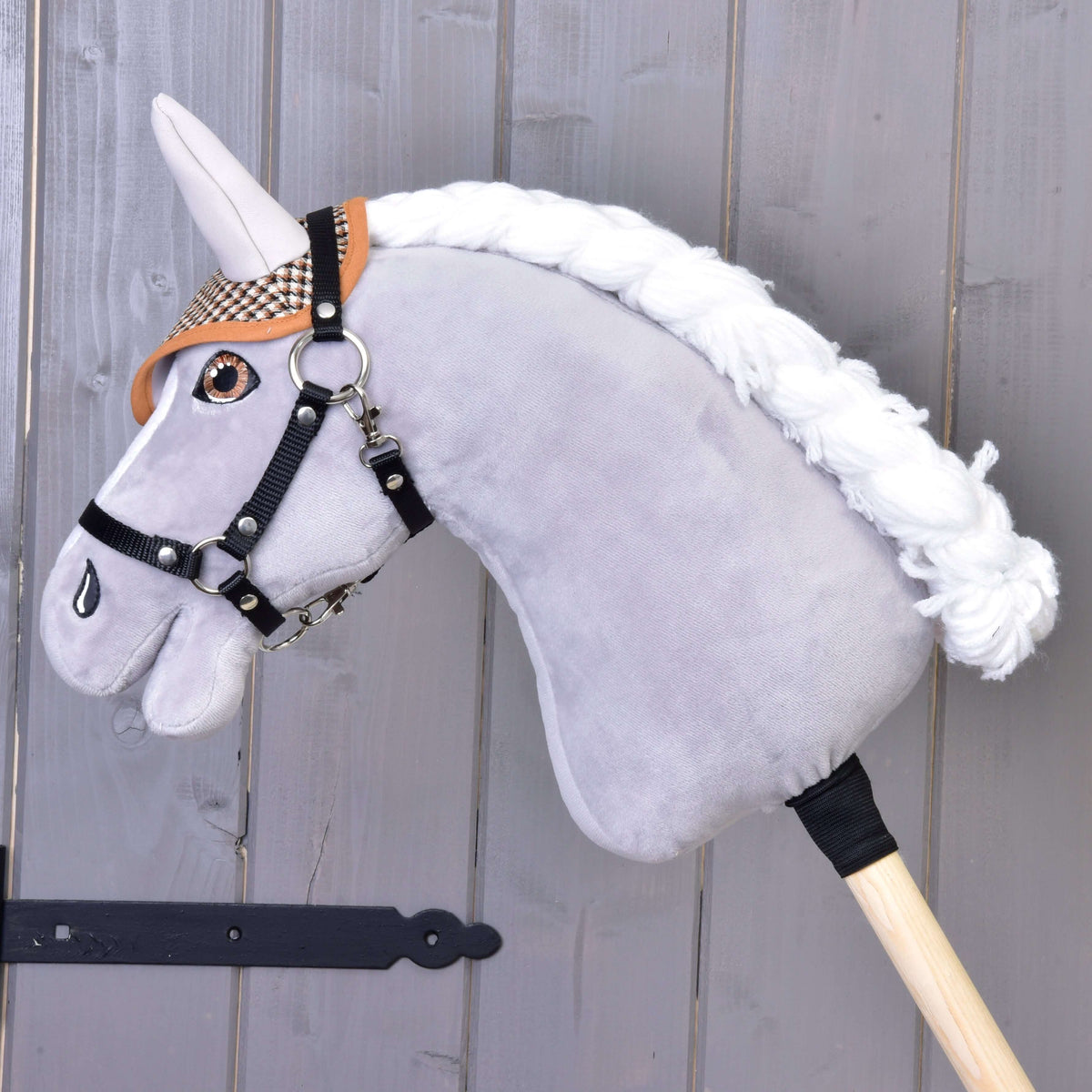 The Hobby Horse LarDen plush hobby horse features a gray head, black bridle, brown plaid "Ear bonnet english brown plaid" for insect protection, white mane, and is mounted on a wooden stick against a wood panel background.