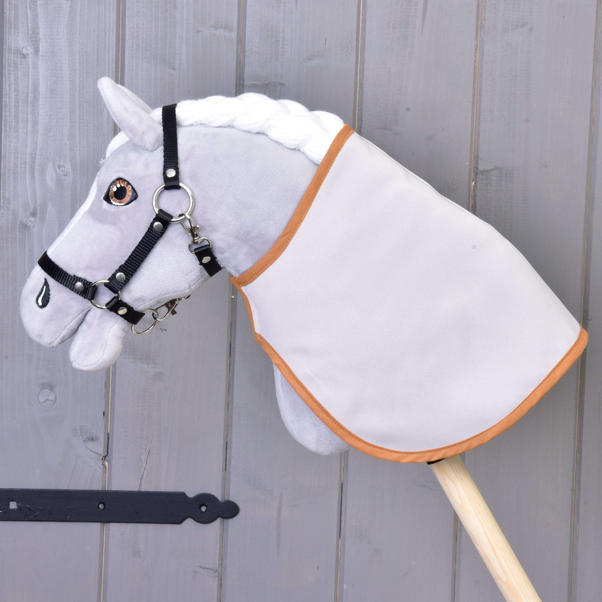 A gray Hobby Horse LarDen with a white mane, black bridle, and a Blanket english brown plaid double-sided is mounted on a wooden stick in front of a gray wooden wall.