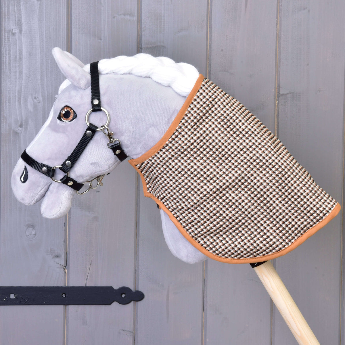 A Hobby Horse LarDen plush hobby horse with a white mane, black bridle, and a Blanket english brown plaid double-sided is mounted on a wooden stick, set against a gray wooden wall.