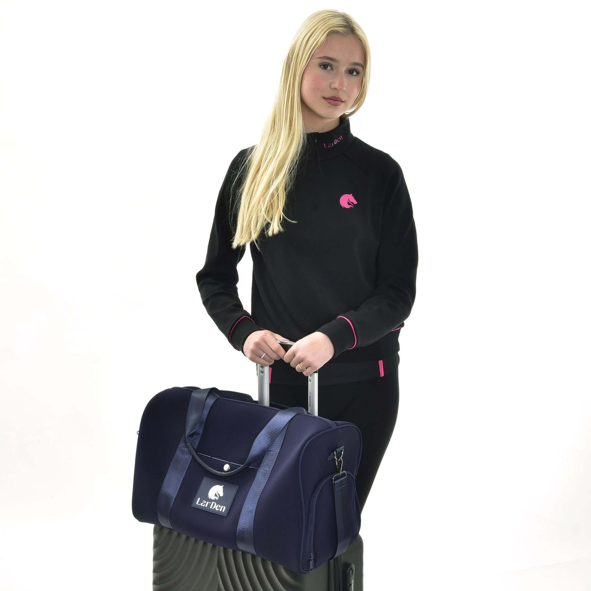 Blonde girl in a black sweatshirt with pink details holding a suitcase with the navy Hobby Horse Sports bag on top
