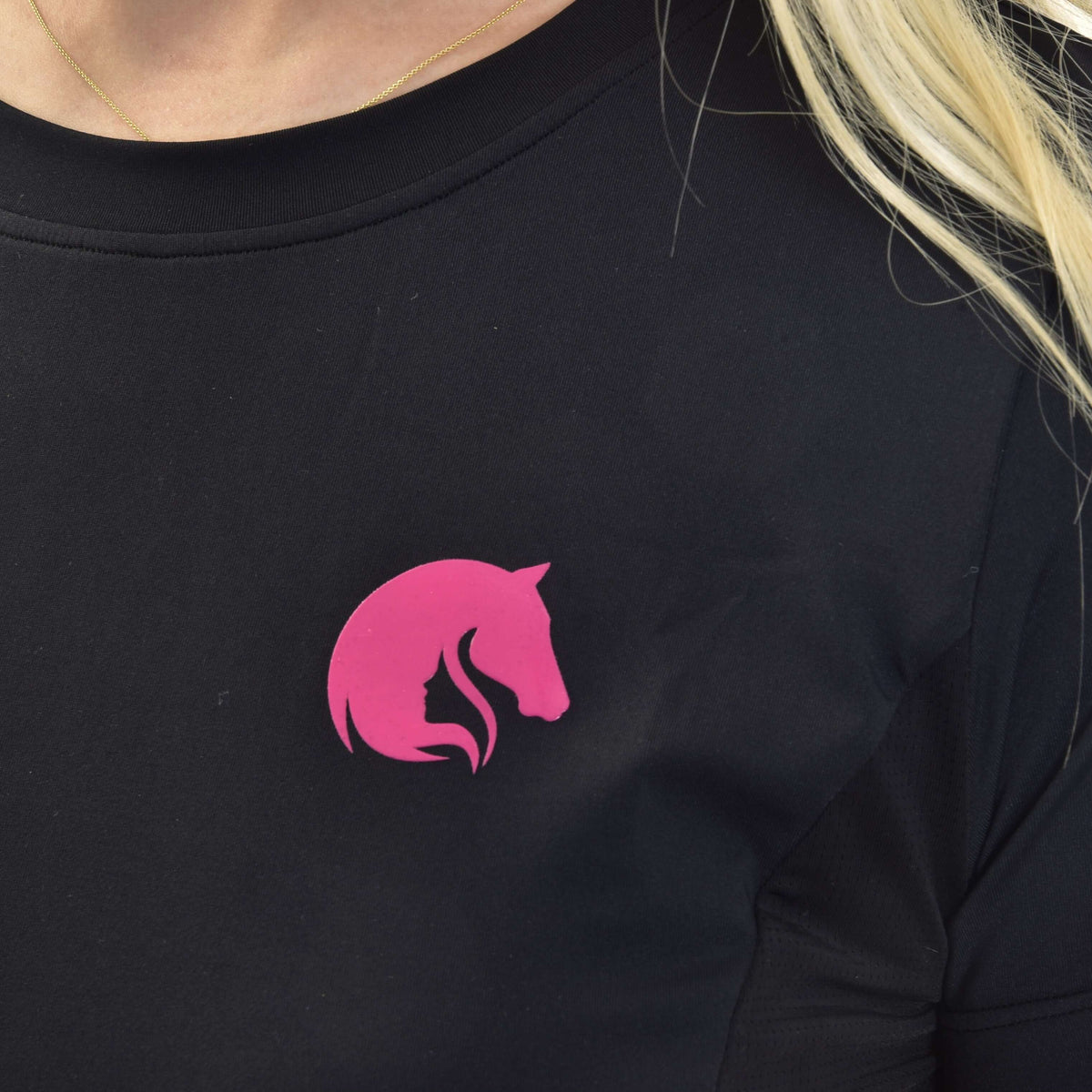 Detalje af det lyserøde hestehovedlogo på en sort sportstrøje med rund hals, båret med en tynd guldkæde