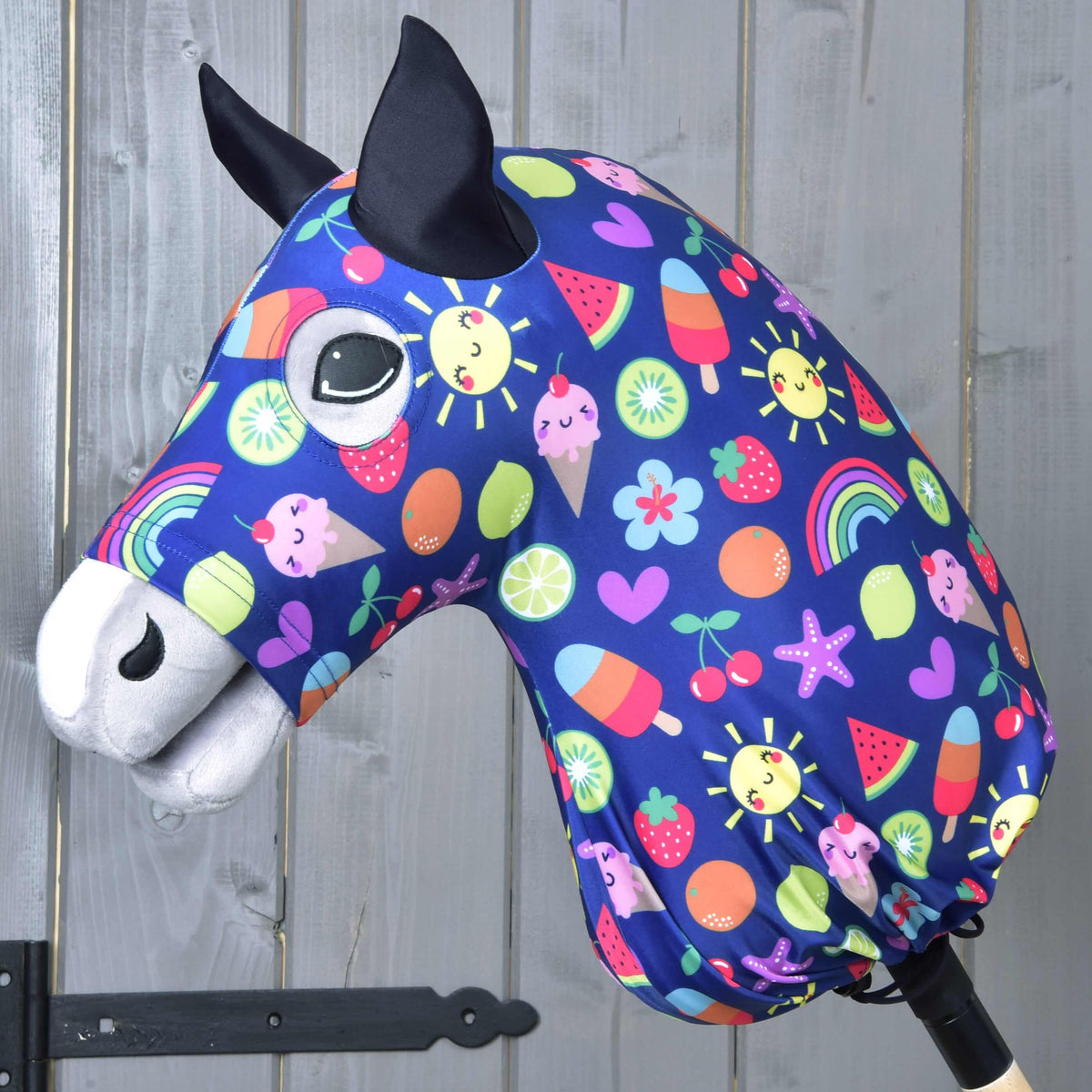 A fabric Hobby Horse head with black ears, large eyes, and a blue Hobby Horse LarDen “Happiness” protective suit featuring suns, rainbows, hearts, fruit, and ice cream—set against a wooden background.