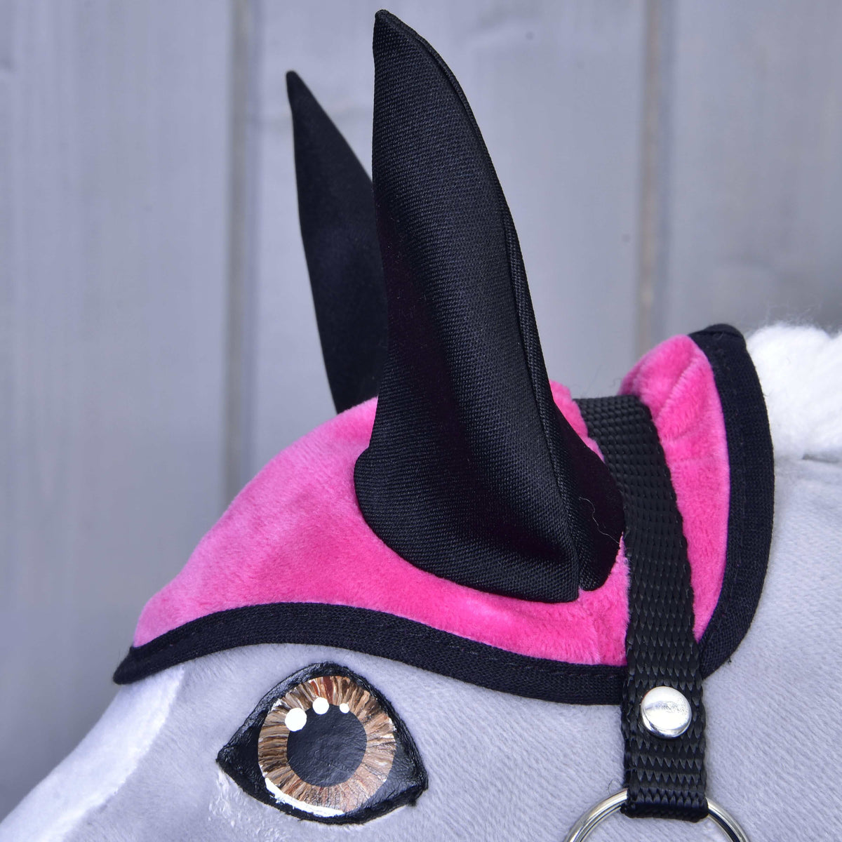 A close-up of a gray toy horse’s head wearing the Set Hot Pink ear bonnet by Hobby Horse LarDen, featuring large fabric ears and a black strap, against a light wooden background—part of the ear bonnet and blanket set.