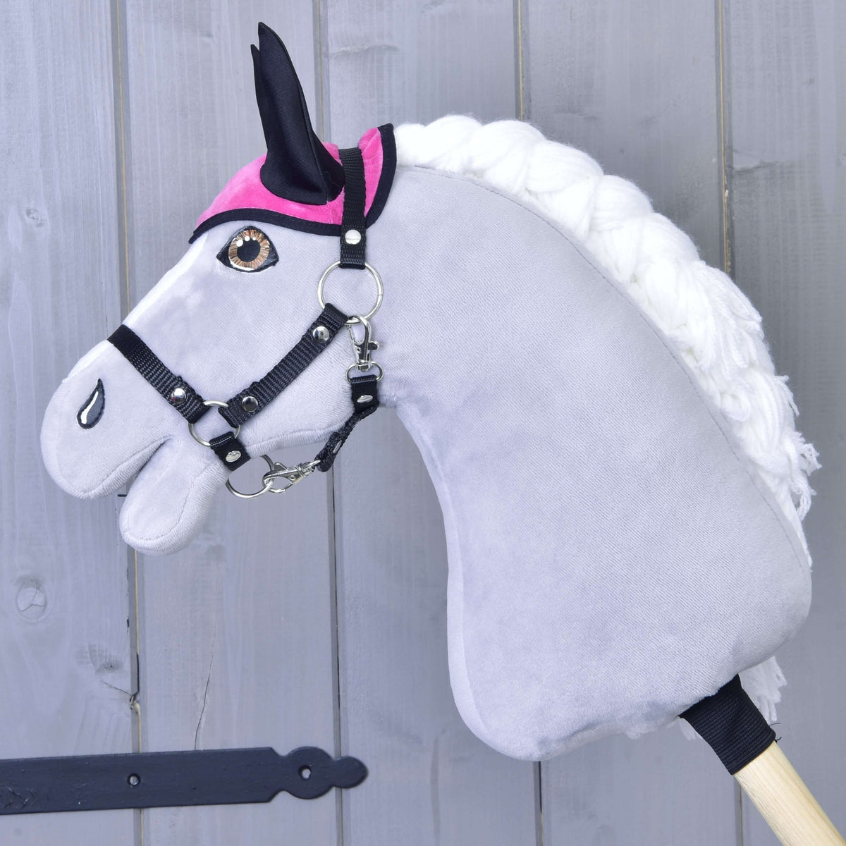 The Hobby Horse LarDen Set Hot Pink includes an ear bonnet and blanket for your plush grey hobby horse with a white mane, black bridle, and pink accessories, mounted on a wooden stick in front of a light grey wood wall.