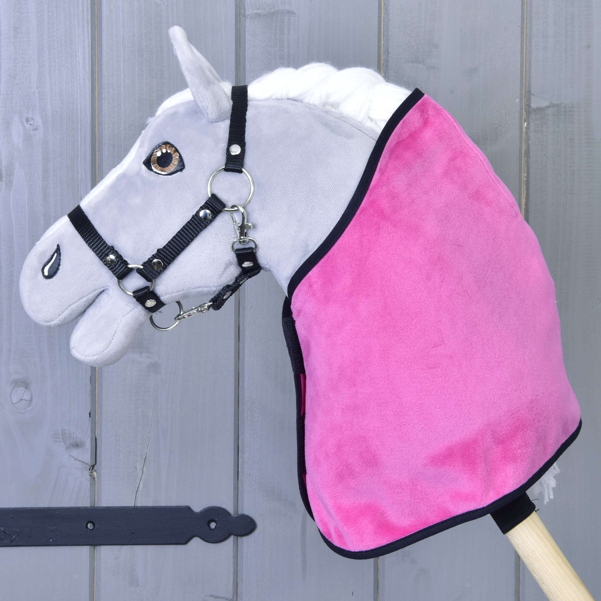 The Hobby Horse LarDen Set Hot Pink includes a gray hobby horse with black bridle, hot pink blanket, and matching ear bonnet on a wooden stick, displayed before a rustic gray wooden wall.