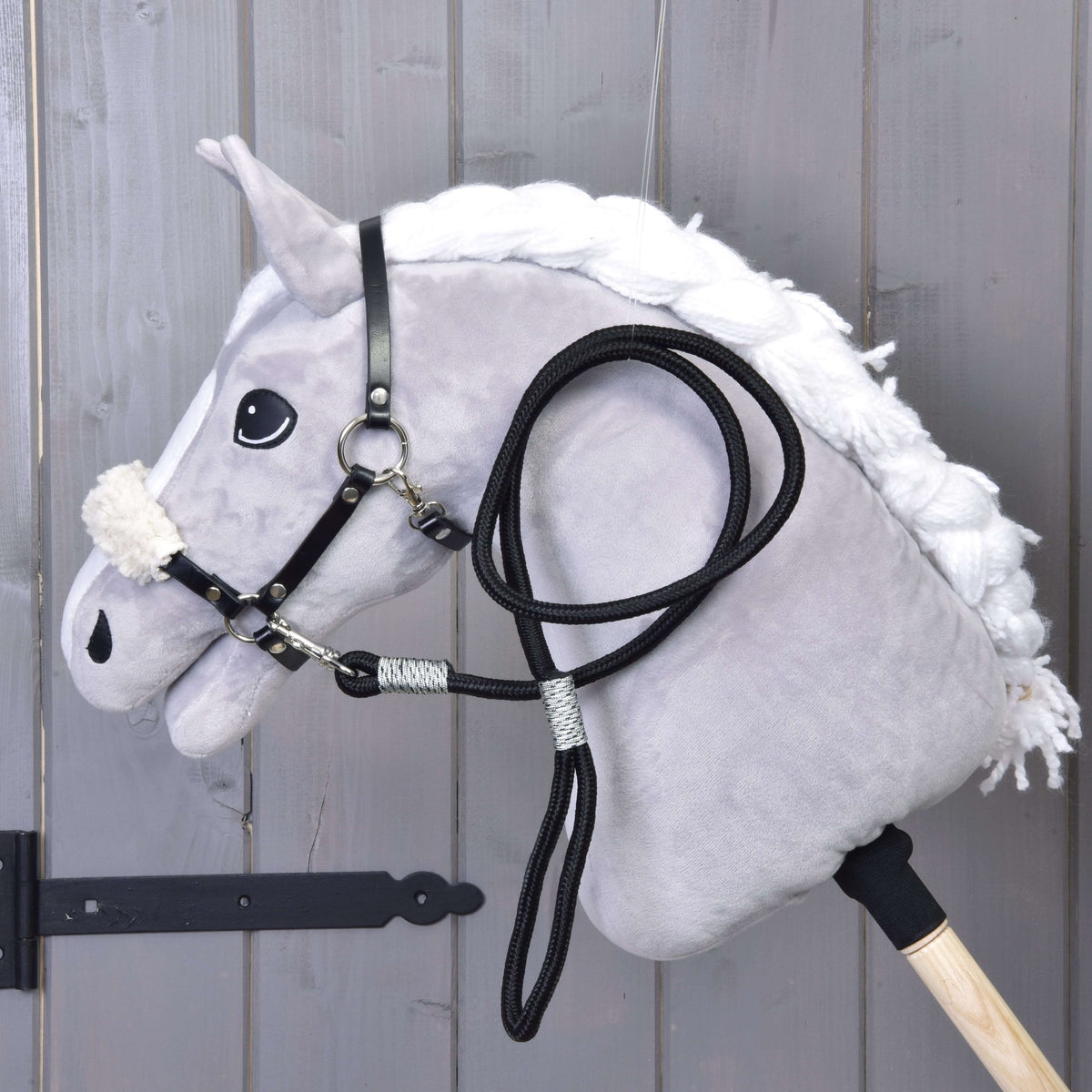 The Hobby Horse LarDen Set Basic features a plush hobby horse with a white mane, black bridle with reins, and black halter on a wooden stick, shown leaning against gray wooden panels.