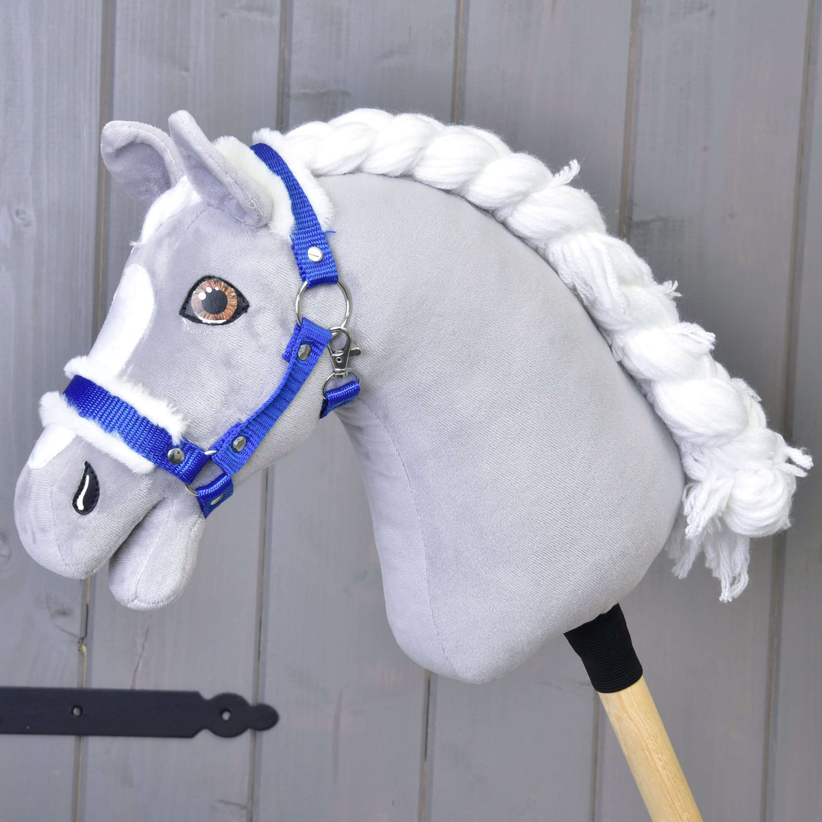 A gray fabric hobby horse with a white yarn mane and the "Hobby Horse LarDen" Halter blue with fur stands in front of a light gray wooden wall.