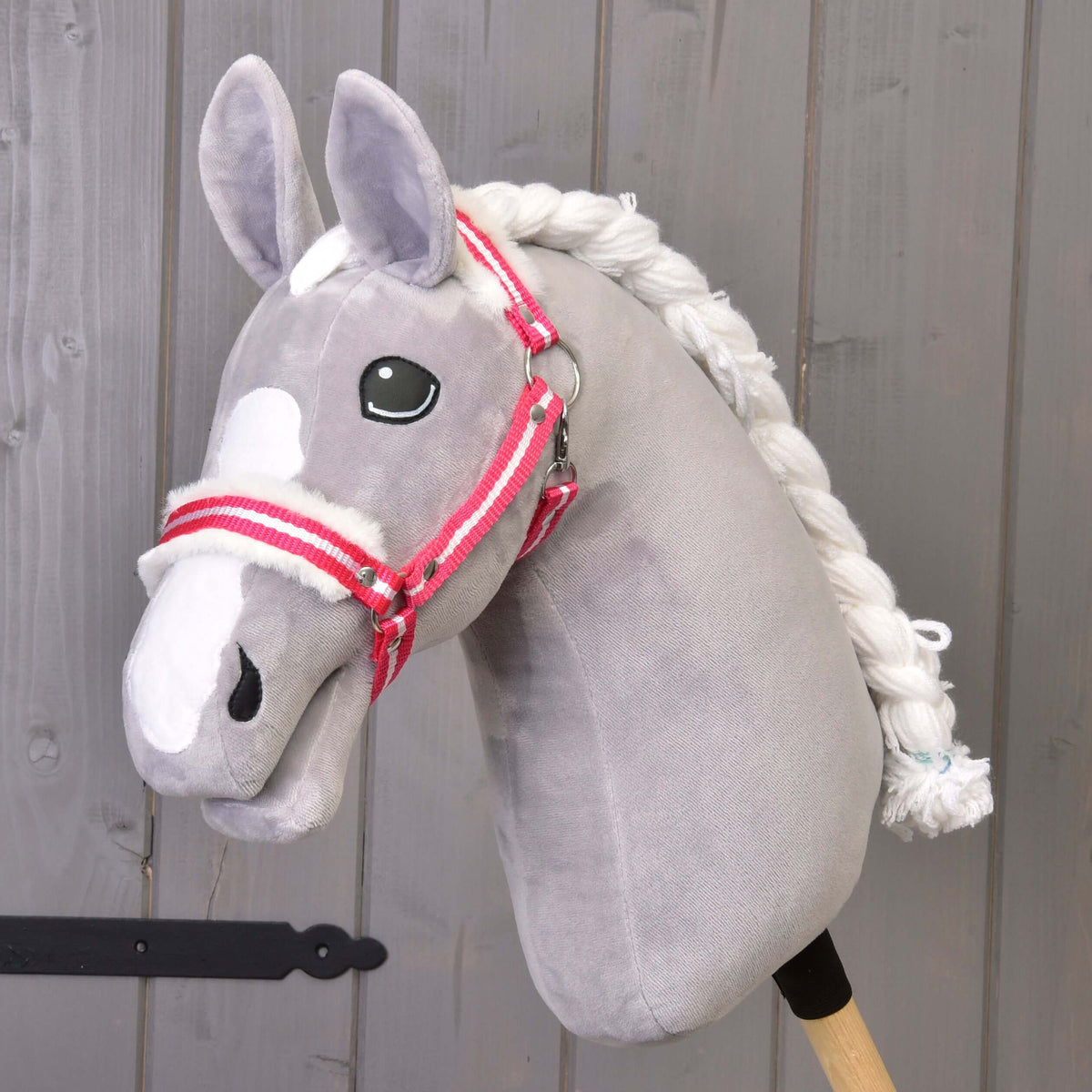 The Hobby Horse LarDen "Halter red stripe with fur" is a gray plush hobby horse with a white mane and red stripe halter on a wooden stick, shown in front of a gray wooden wall.
