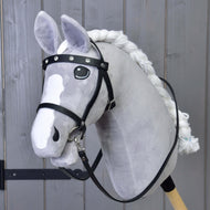 A gray plush hobby horse by Hobby Horse LarDen features the Complete black bridle with diamonds, white mane, detachable reins, and wooden stick, posed before a gray wooden wall.