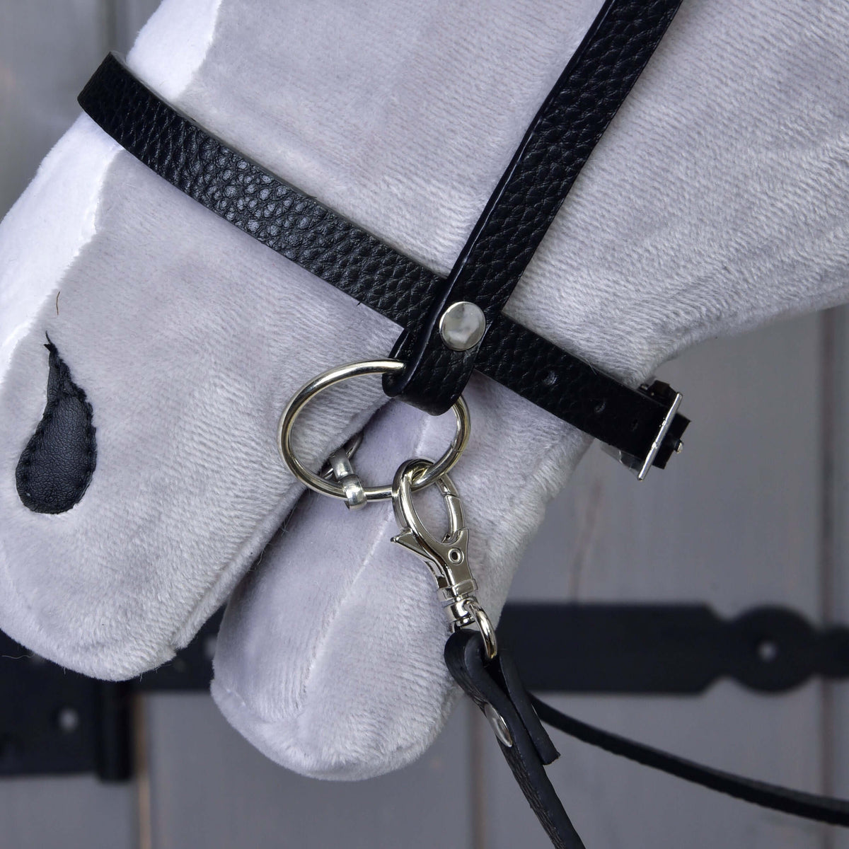 A close-up of a white plush horse’s head showcases the Hobby Horse LarDen Complete black bridle with diamonds, featuring detachable reins and an English noseband with metal bit, set against a wooden background with visible dark hardware.