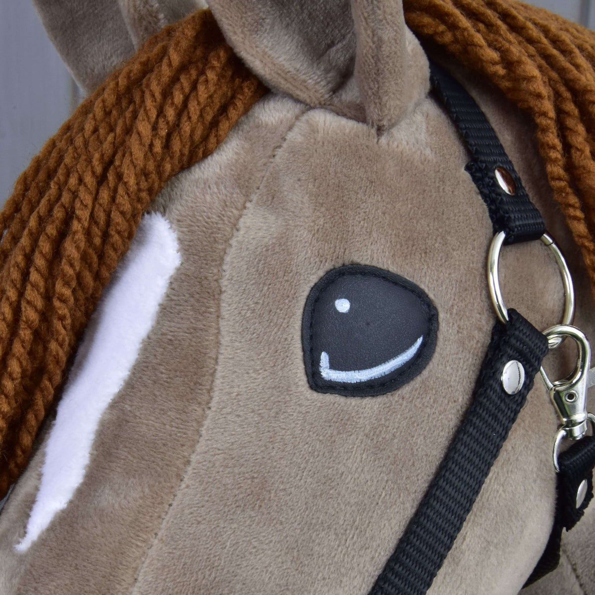 Close-up of Hobby Horse LarDen’s “Hobby Horse Oliver” with a black halter (plus free fur halter), featuring a brown braided yarn mane, white eye patch, and cartoon-style sewn-on eye—adorable dressage horse look.
