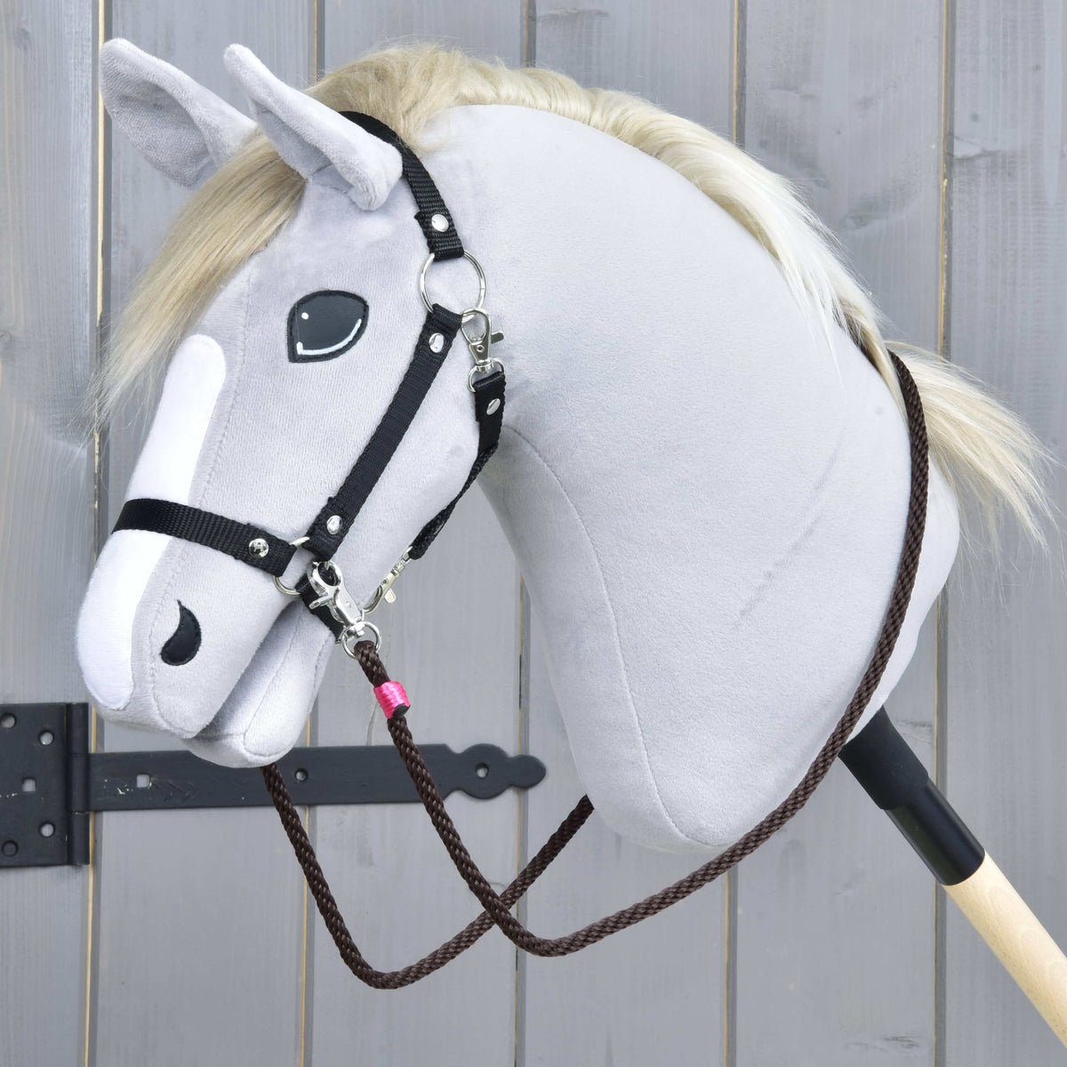 A close-up of the Rope set Noir Cocoa by Hobby Horse LarDen—a gray plush hobby horse with black bridle, halter, and brown reins on a wooden stick—set against a light wooden background.