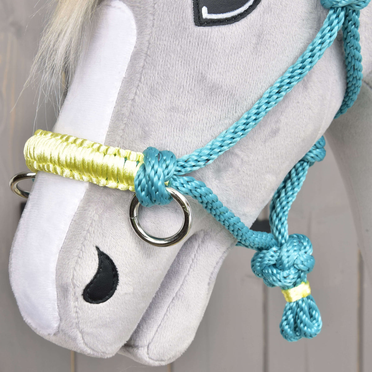 A close-up of a gray plush hobby horse toy by Hobby Horse LarDen, featuring a Rope halter Turquoise and bridle, black stitched eyes and nostrils, and a mane made of white yarn.