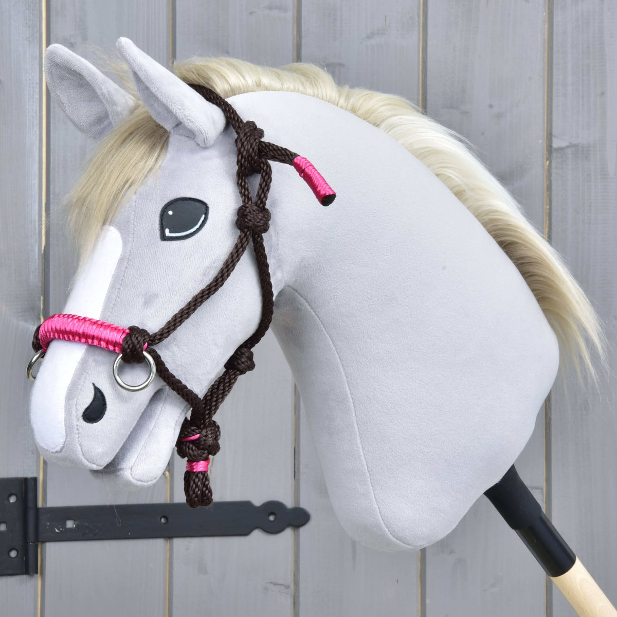 The Hobby Horse LarDen Rope set Noir Cocoa is a gray plush hobby horse on a stick with a black and pink bridle, reins, black button eyes, and a white mane, shown in front of a wooden fence.