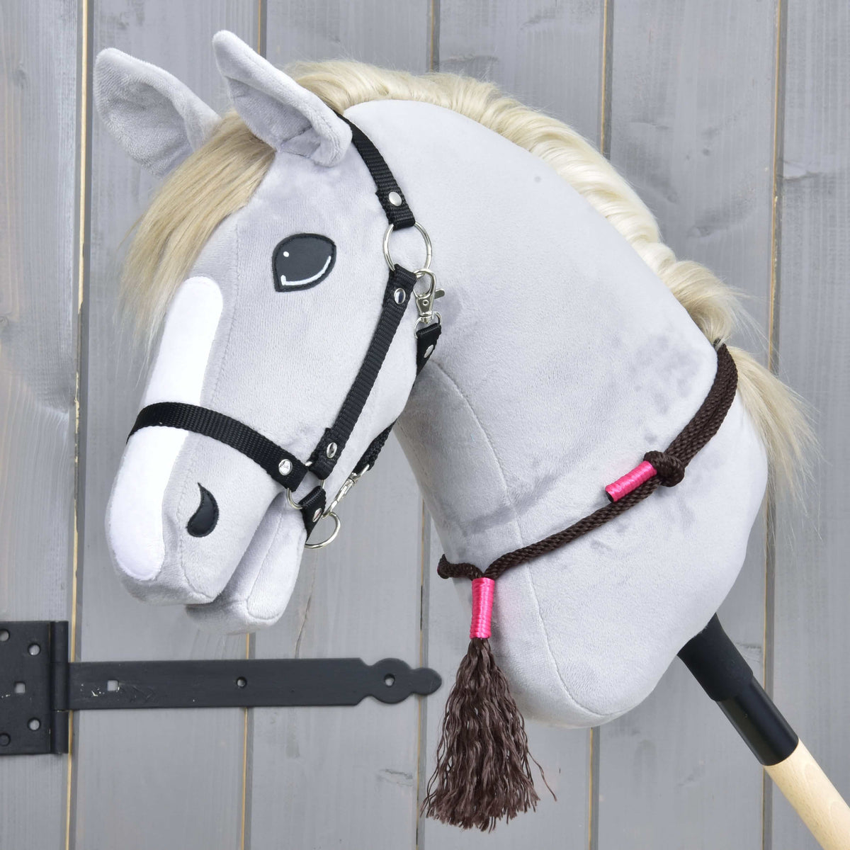 The Hobby Horse LarDen Rope set Noir Cocoa, a gray plush hobby horse with a white mane, black bridle, and brown rope reins with pink accents on a wooden stick, rests against a gray wooden background.