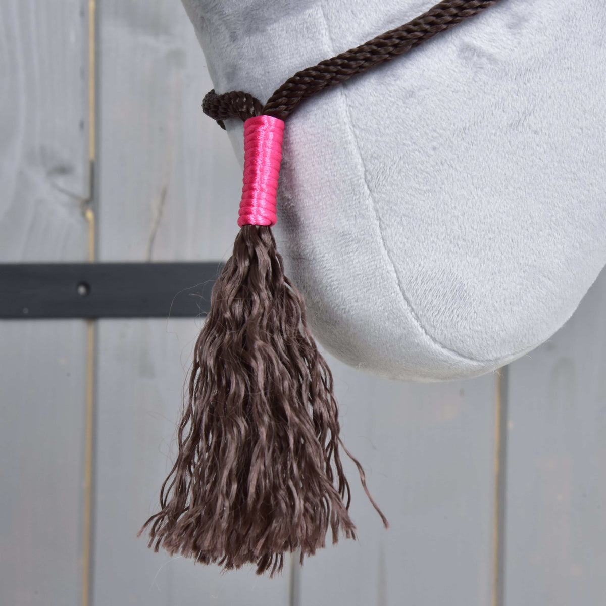A close-up of the Rope set Noir Cocoa by Hobby Horse LarDen shows a braided dark brown tassel on thick reins with a pink wrap, displayed against a light gray plush surface and wooden background.
