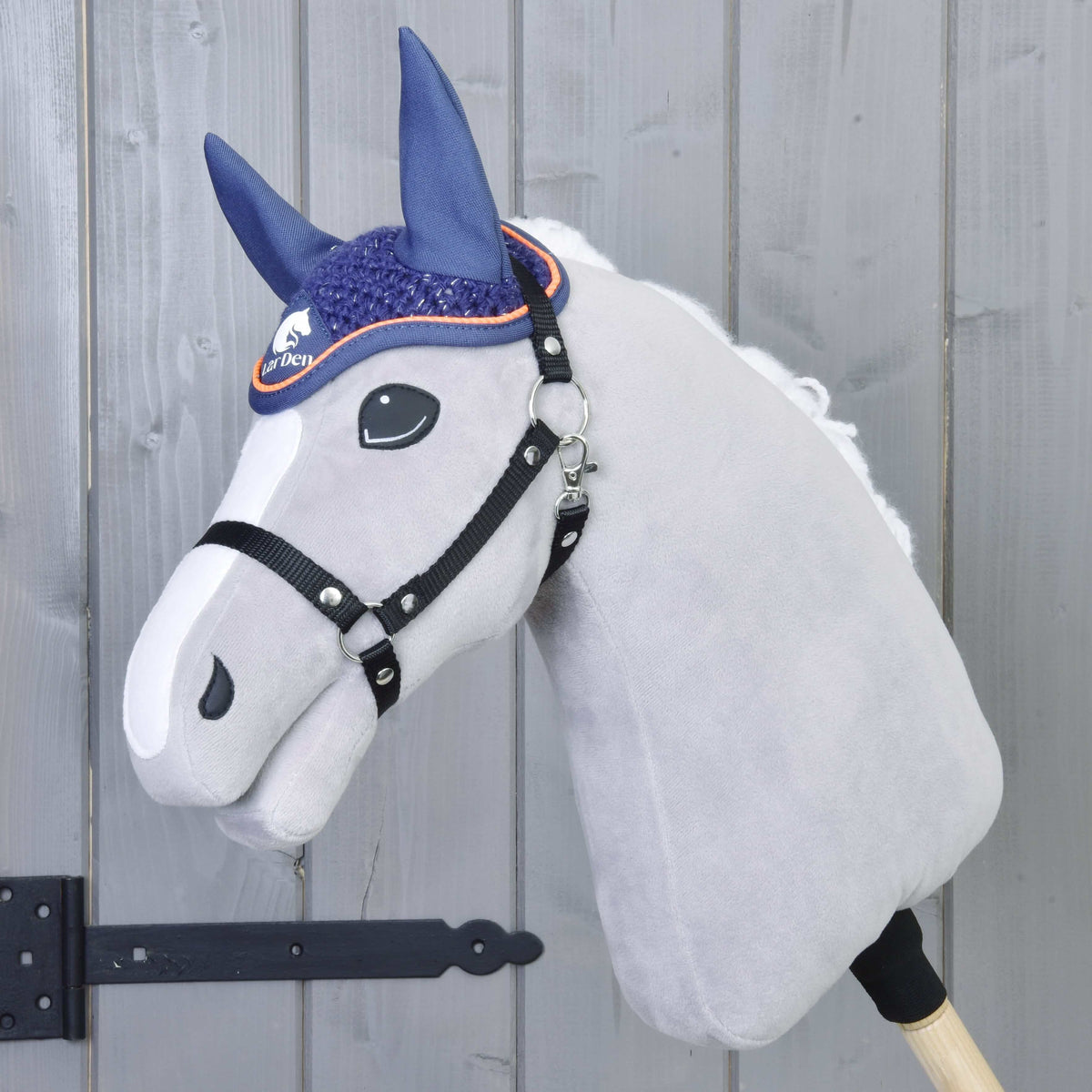 The Hobby Horse LarDen Ear Bonnet Crocheted Blue Sport is shown on a plush hobby horse with a white face and black bridle, mounted on a wooden stick in front of a gray panel door with a black hinge.
