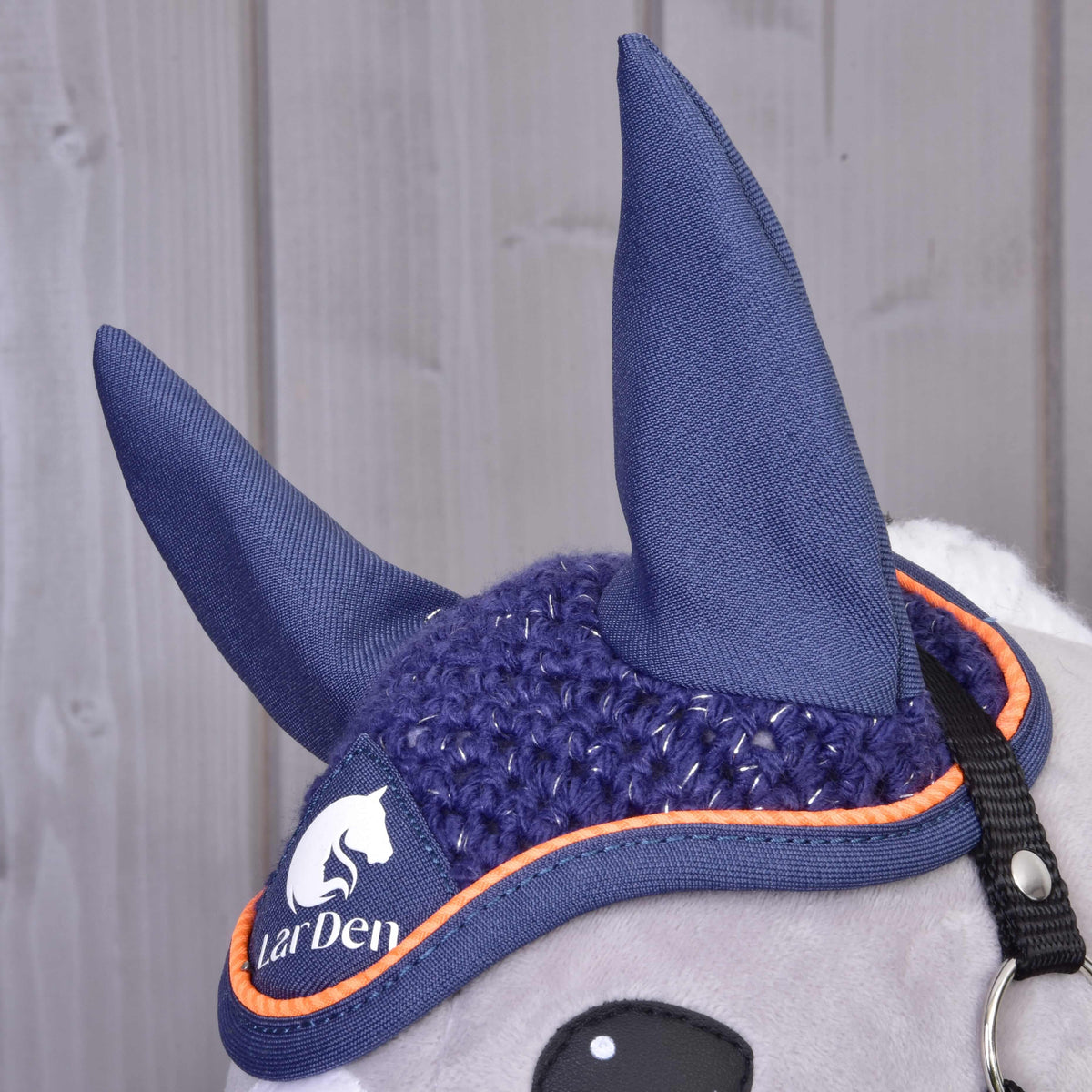 A close-up of a stuffed horse toy’s head wearing the Hobby Horse LarDen Ear bonnet crocheted Blue Sport with orange trim and logo. Its soft grey fabric, black bridle, and the stylish bonnet offer insect protection against a wooden background.