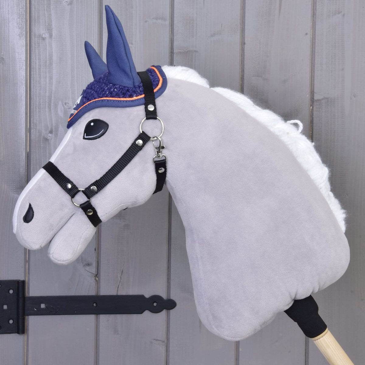 The Hobby Horse LarDen "Ear bonnet crocheted Blue Sport" features a plush light gray head, white mane, black bridle, and a crocheted blue ear bonnet on a wooden stick, shown against a light wooden wall.