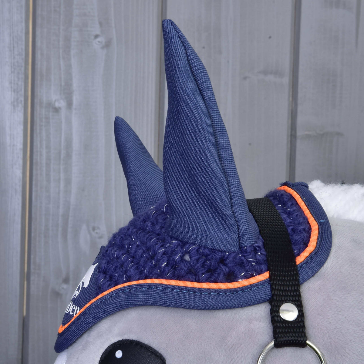 Close-up of the Hobby Horse LarDen Ear bonnet crocheted Blue Sport on a toy horse’s head. This blue ear bonnet with orange trim and black strap features two upright ears and protects hobby horses from insects.