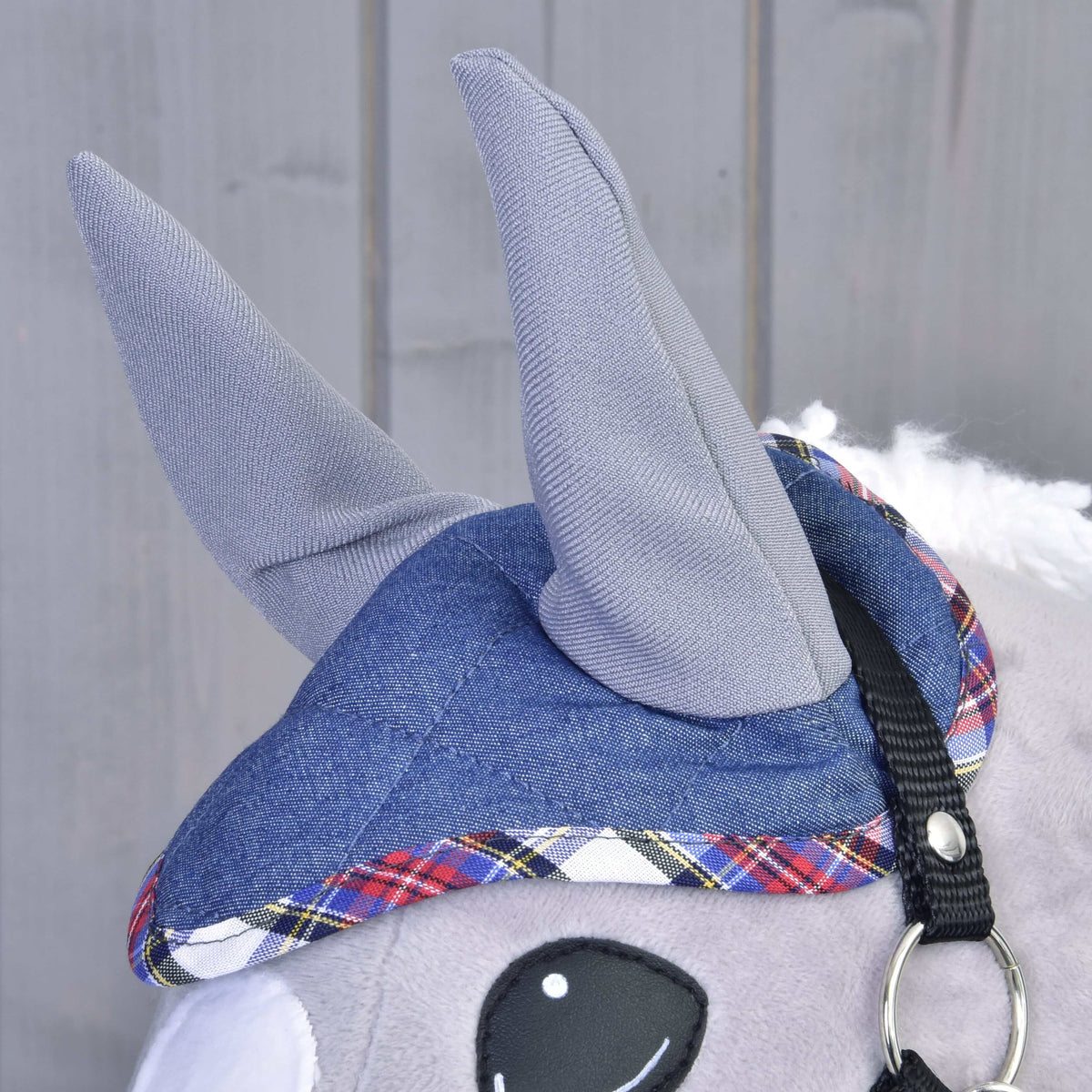 Close-up of the Set Jeans plush animal by Hobby Horse LarDen, showing gray fabric ears, a blue plaid hat like an ear bonnet set, a black strap with metal ring, and a large white and black eye against a wooden background.