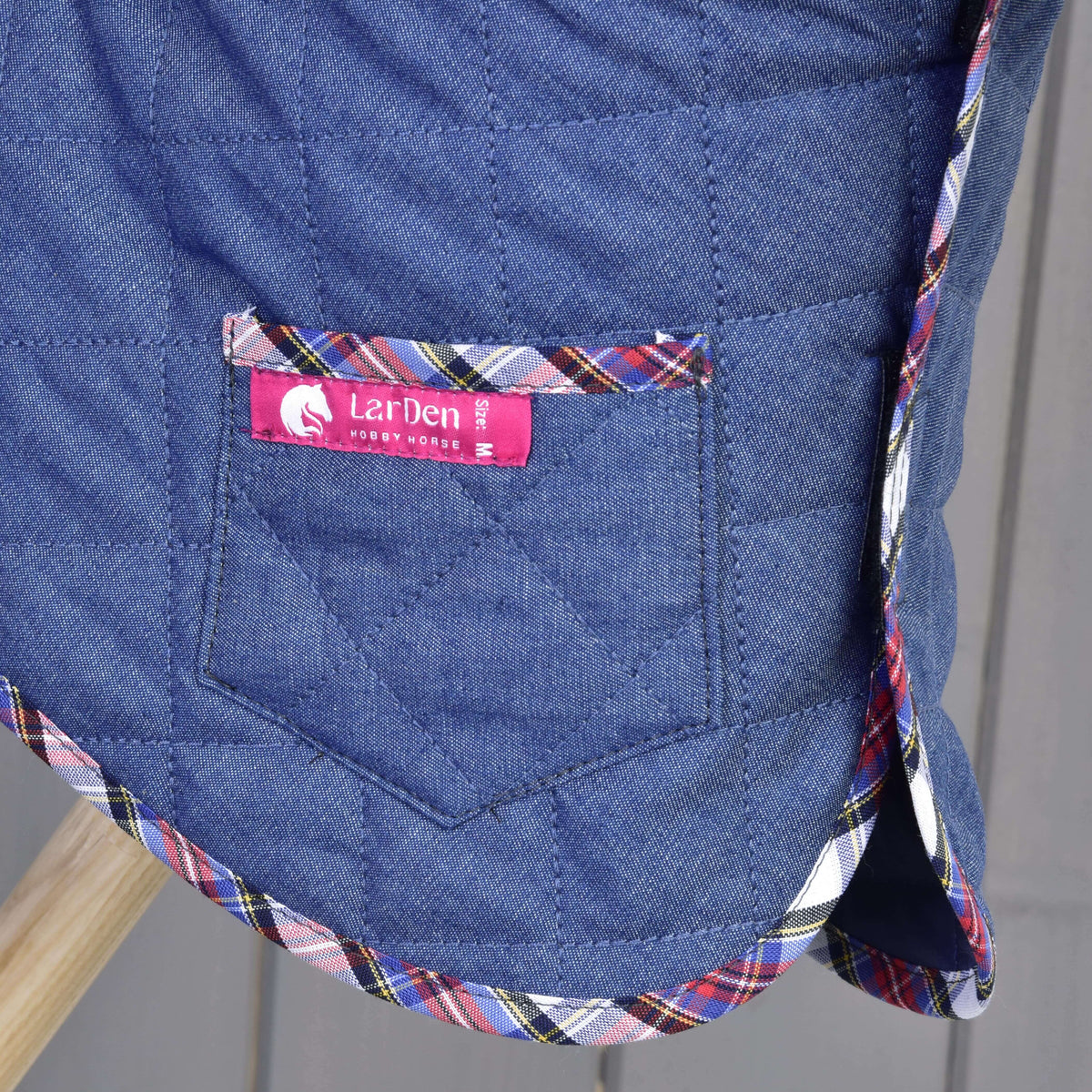 A close-up of Set Jeans, a blue quilted blanket by Hobby Horse LarDen, featuring a pocket, multicolored plaid trim, and a pink label that says Hobby Horse LarDen.