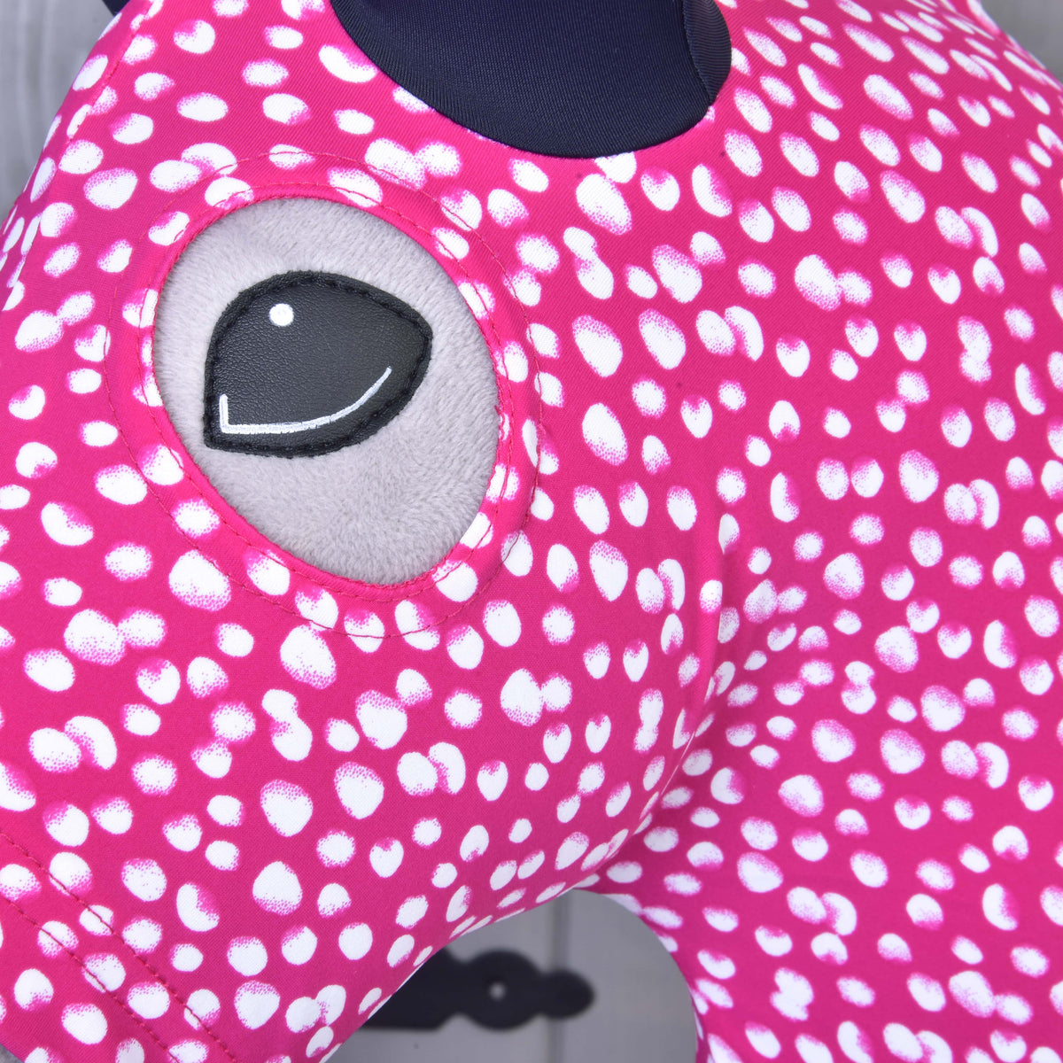 Close-up of the Hobby Horse LarDen Hobby Horse Protective suit Minie (Lycra), a pink, white-spotted washable cover with an embroidered gray eye. Keeps your Hobby Horse clean and stylish. Blurred background shows part of a gray wall.