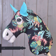 The Hobby Horse LarDen’s Protective suit Pineapple (Lycra) features a black head with a vibrant pineapple and leaf print, blue ears, and a white muzzle, mounted on a wooden stick and displayed against a gray wooden wall.