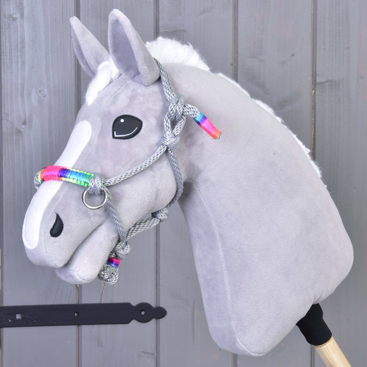 The Hobby Horse LarDen Rope Halter Manhattan—a soft gray hobby horse accessory with a white face patch, black button eyes, and a rainbow-accented braided rope halter—displayed on a gray wooden background.