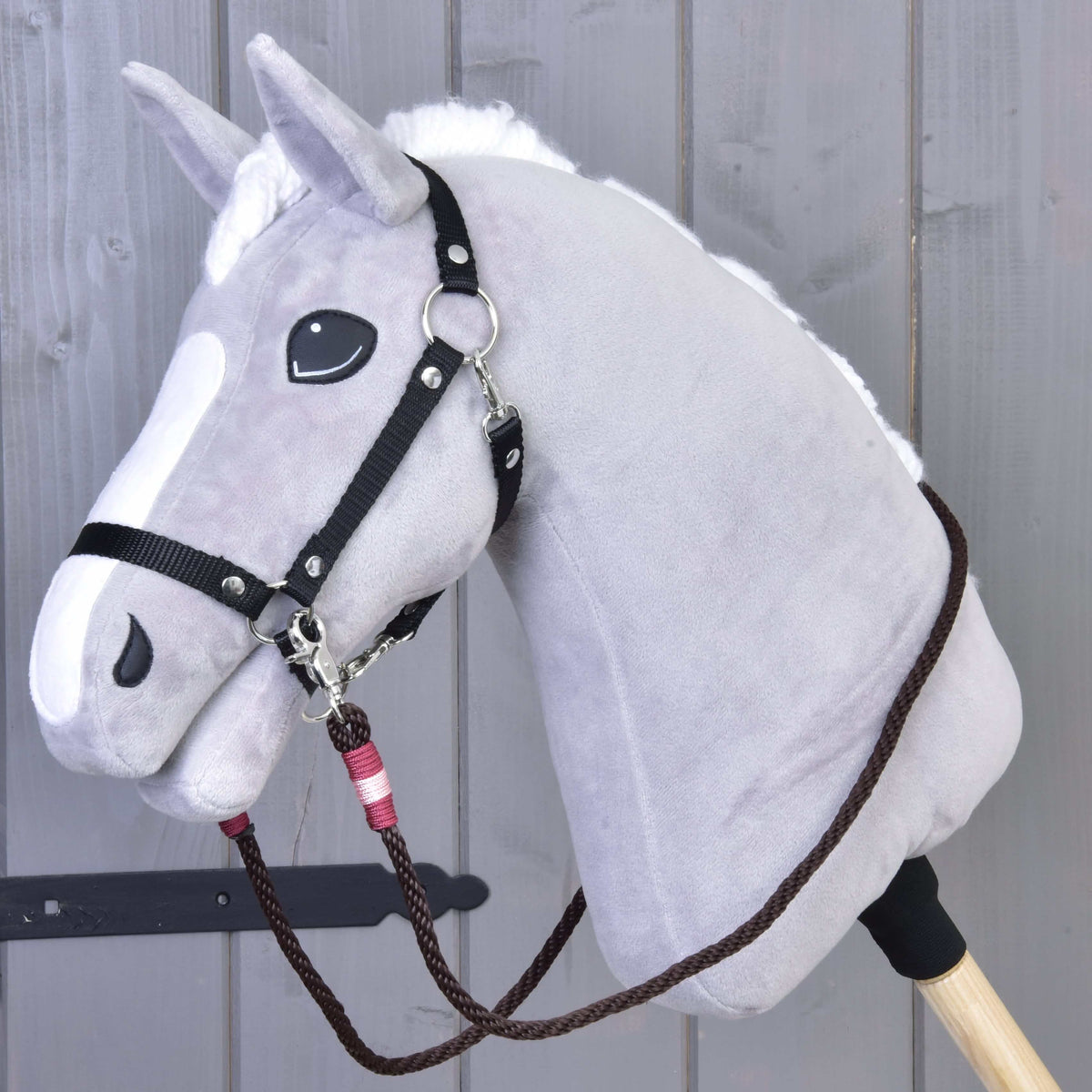 The Hobby Horse LarDen Reins York is a gray plush hobby horse with a white mane and snout, featuring black handmade polypropylene rope reins, mounted on a wooden stick against a wooden panel background.