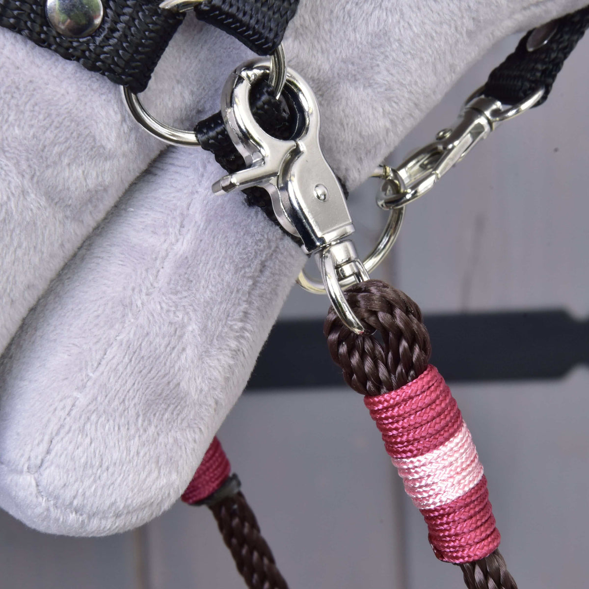 Close-up of a metal clip attaching the Hobby Horse LarDen Reins York—brown and pink braided polypropylene rope—to a black fabric strap on a soft, light gray surface, likely part of hobby horse tack.