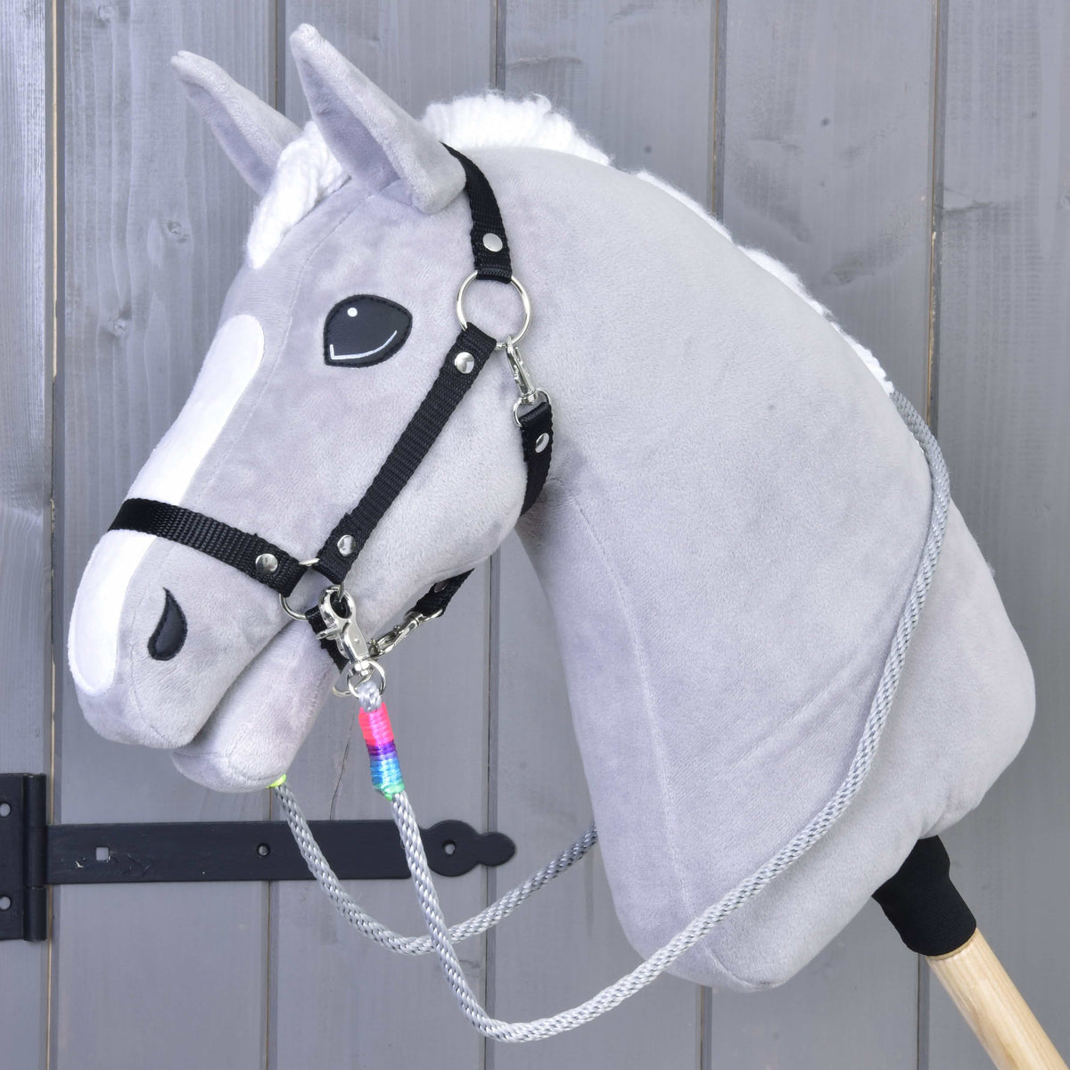 The Hobby Horse LarDen Reins Manhattan features a gray hobby horse with a white mane, black halter, handmade reins, and a wooden stick, shown against a gray wooden wall.