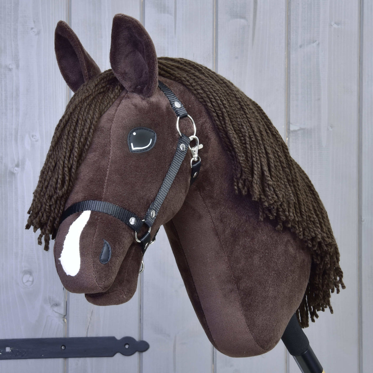 Hobby Horse LarDen’s Chianti, a brown plush hobby horse with black halter, button eyes, yarn mane, and a white nose patch inspired by American Quarter Horses, posed before a light gray wooden background.