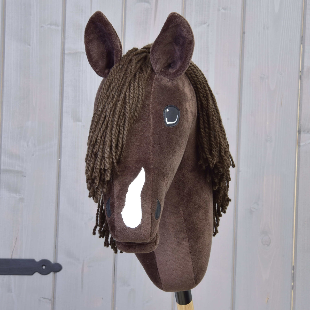 The Hobby Horse LarDen "Hobby Horse Chianti with Black Halter" is a brown plush hobby horse with a white blaze, black button eyes, and a brown yarn mane, inspired by the gentle American Quarter Horse. Set against a wooden panel background.