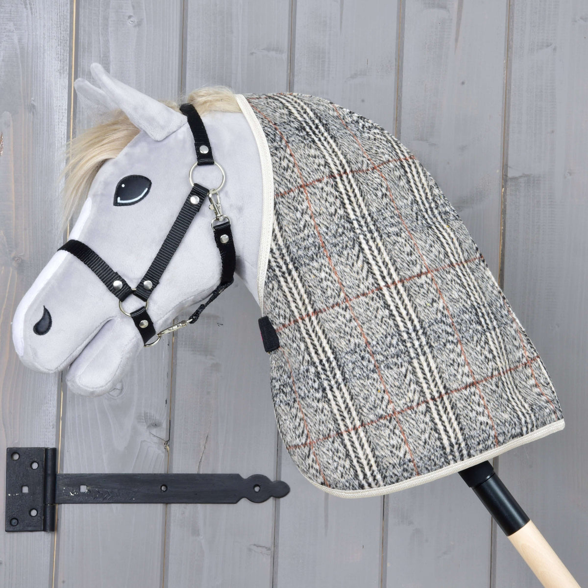 A gray plush hobby horse from Hobby Horse LarDen features a black bridle and the Stable Blanket Fine Check, mounted on a wooden stick and set against a light gray wooden wall.