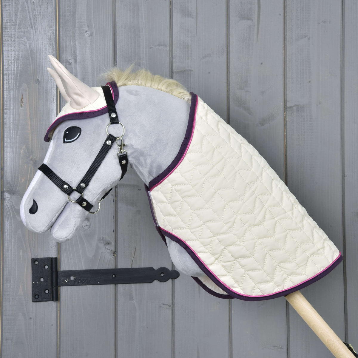 The Set Supreme Crema by Hobby Horse LarDen features a gray hobby horse with a black bridle, cream quilted blanket with purple trim, matching ear bonnet, and is mounted on a wooden pole against a gray wooden wall.