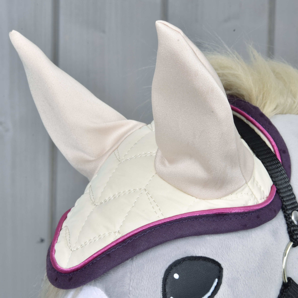 Close-up of a Hobby Horse LarDen Set Supreme Crema: the horse's head wears a cream-colored quilted ear bonnet with large fabric ears and purple trim, set against light gray wooden panels.