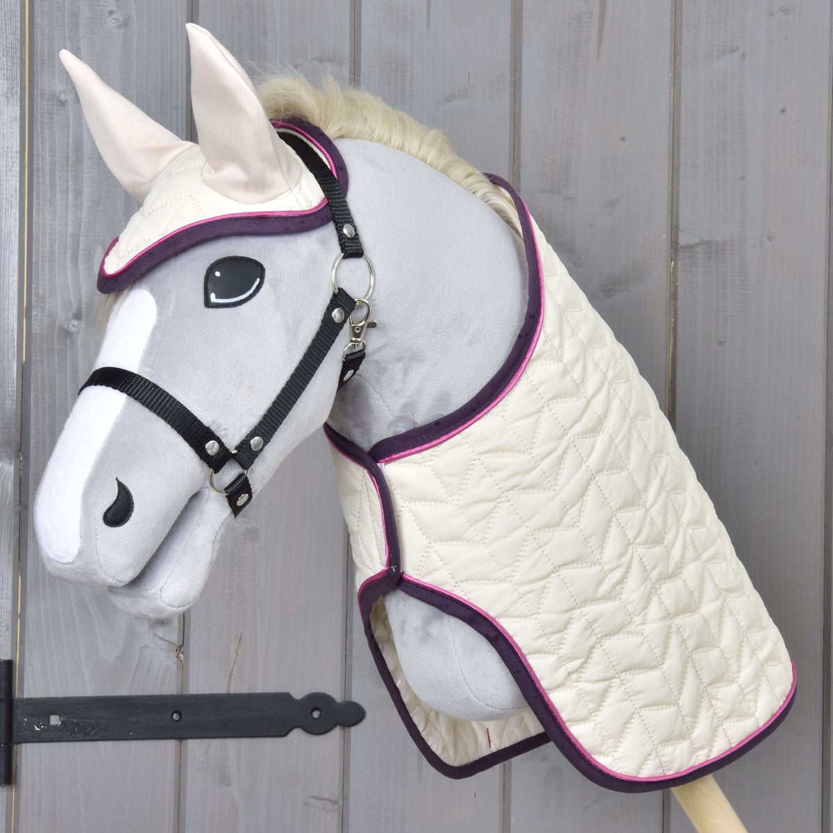 The Hobby Horse LarDen Set Supreme Crema features a plush gray-faced hobby horse with a white muzzle and ears, dressed in a quilted cream blanket and matching ear bonnet with purple trim, displayed on a wooden wall.
