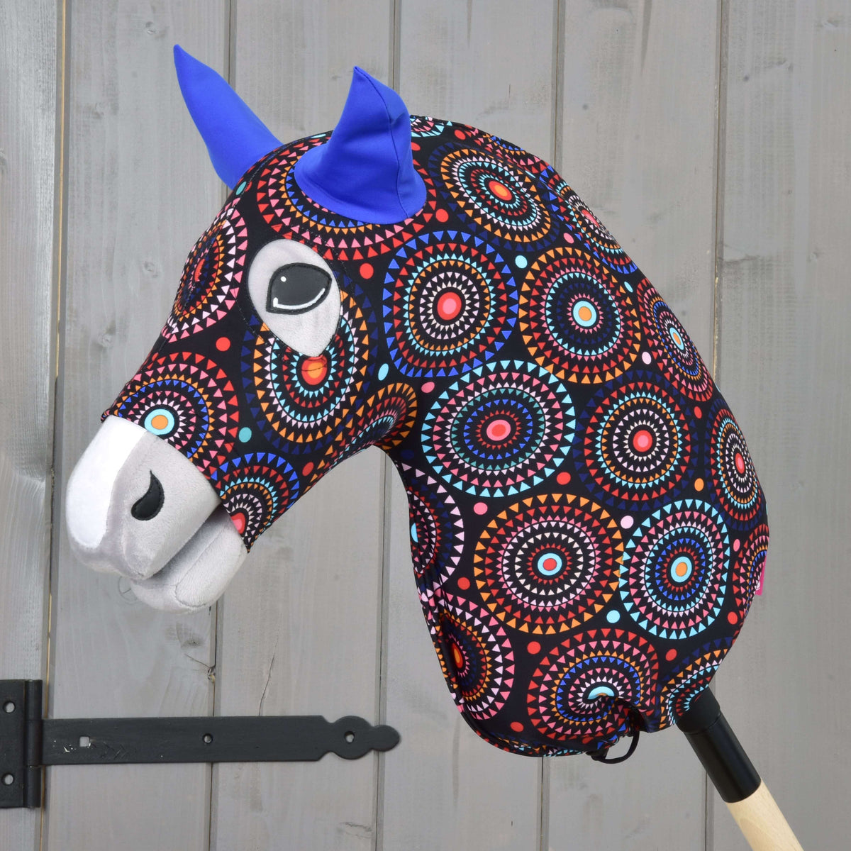 The Hobby Horse LarDen Protective Suit Alice (Lycra) features a colorful patterned head, blue felt ears, and expressive eyes on a wooden stick, displayed against a light gray wooden wall.